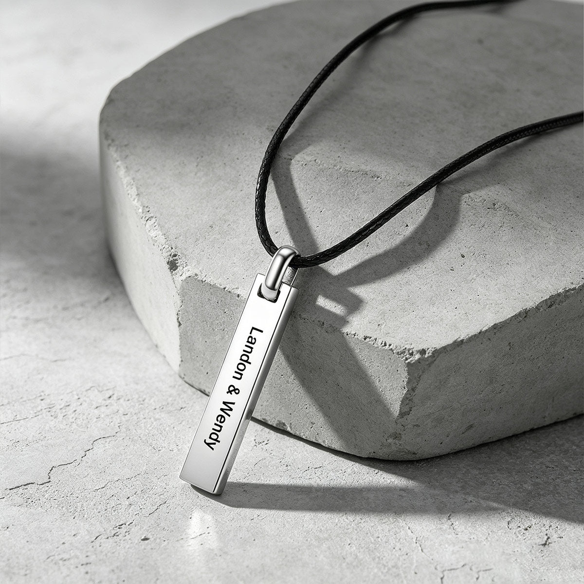 A silver rectangular bar pendant necklace engraved with the names 'London & Wendy' in black lettering, hanging from a black cord on a textured gray stone prop.