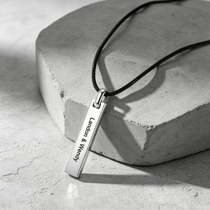 A silver rectangular bar pendant necklace engraved with the names 'London & Wendy' in black lettering, hanging from a black cord on a textured gray stone prop.
