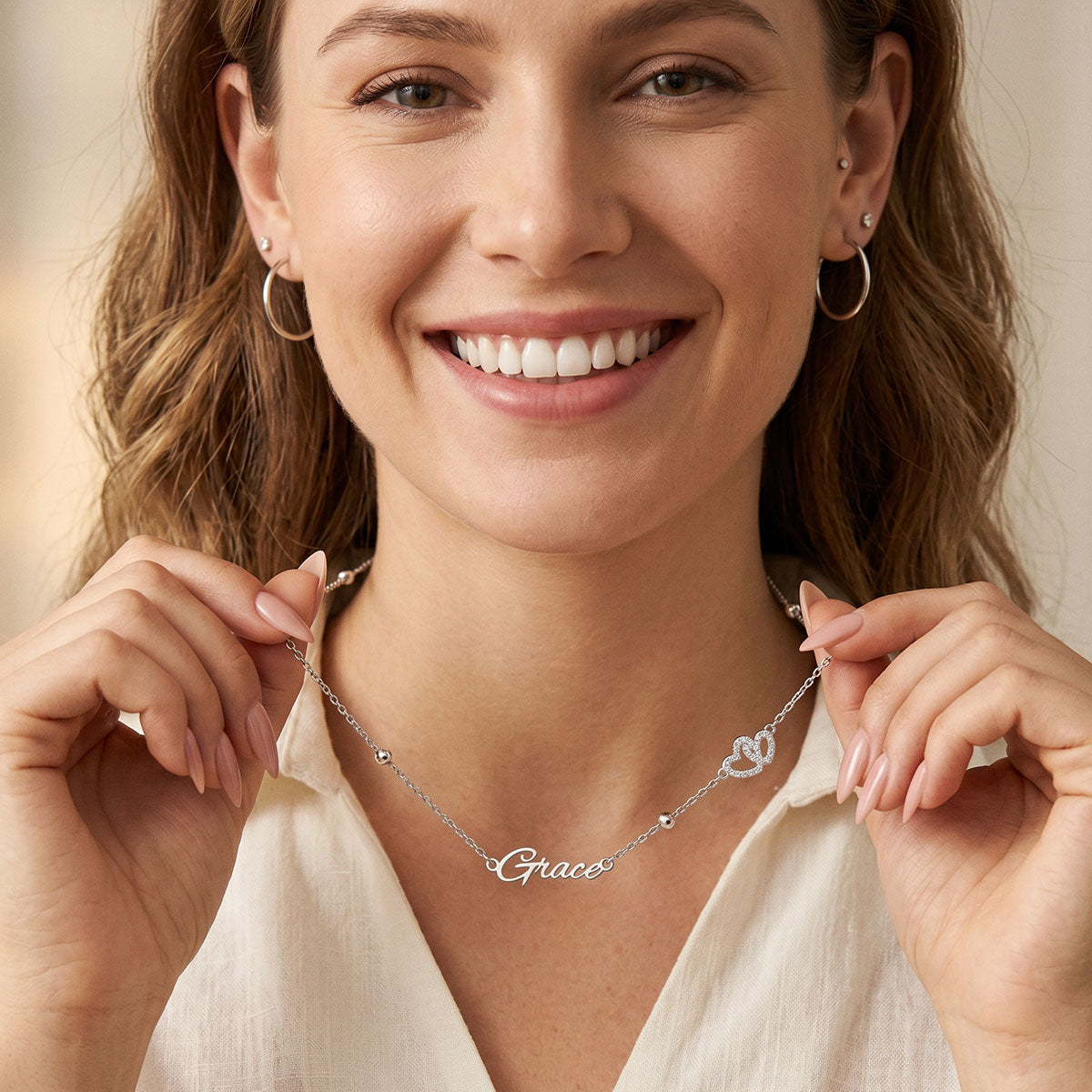 A smiling woman wearing a delicate silver name necklace that reads 'Grace' and a small heart-shaped locket, holding the necklace with both hands against a soft beige background.