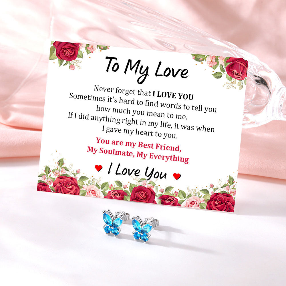 A square white card with floral rose borders and a sentimental love message, styled on a soft pink satin fabric next to a clear glass tumbler. In front of the card are two silver butterfly stud earrings with bright blue rhinestones.