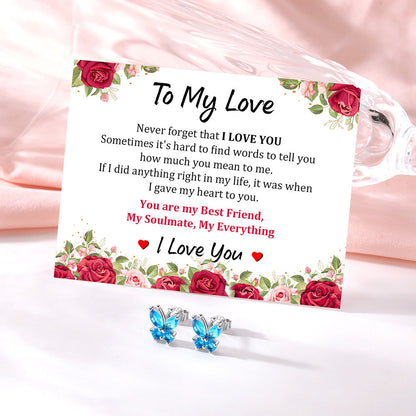 A square white card with floral rose borders and a sentimental love message, styled on a soft pink satin fabric next to a clear glass tumbler. In front of the card are two silver butterfly stud earrings with bright blue rhinestones.