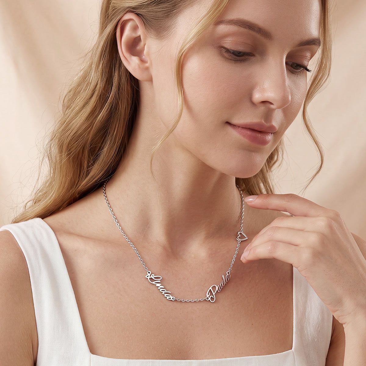 A woman wearing a delicate silver script name necklace with a small heart pendant, styled with a white top against a soft beige background.