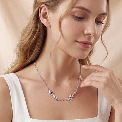 A woman wearing a delicate silver script name necklace with a small heart pendant, styled with a white top against a soft beige background.