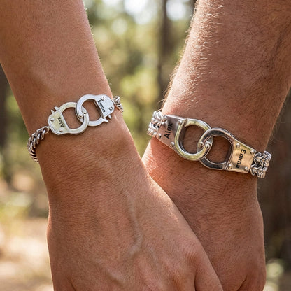 Two wrists wearing handcuff-shaped bracelets with engraved names against a blurred natural background.