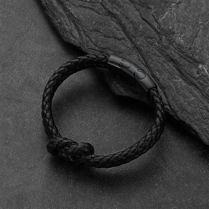 Black braided leather knot bracelet with a matte black metal clasp, displayed on a dark textured slate surface next to its clasp component.