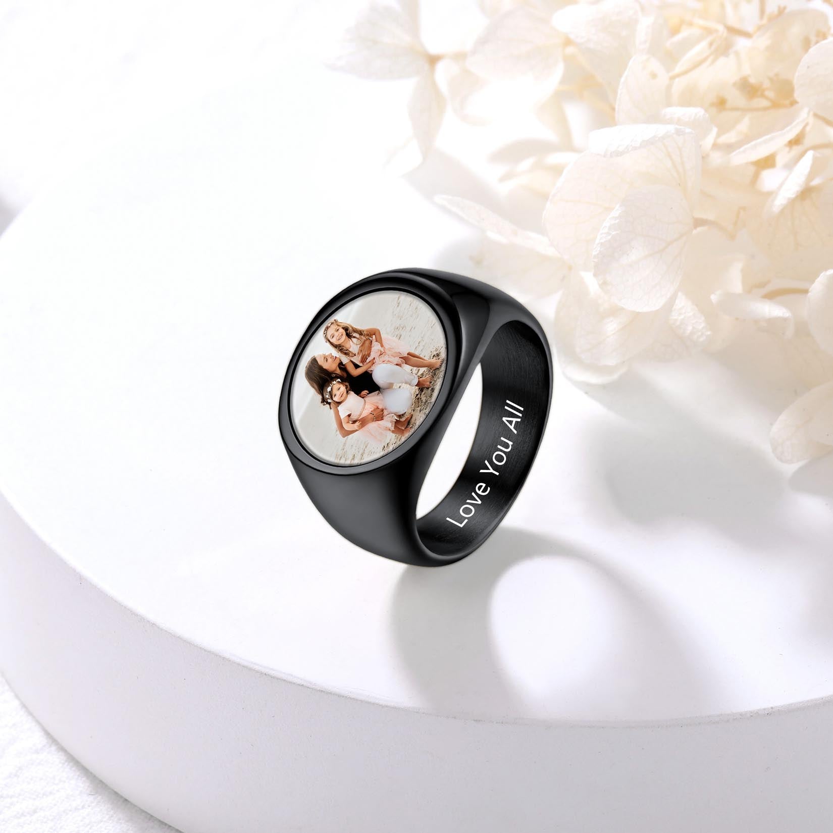 Black picture ignet ring with a photo of two people and 'Love You All' engraving on a white surface with flowers.