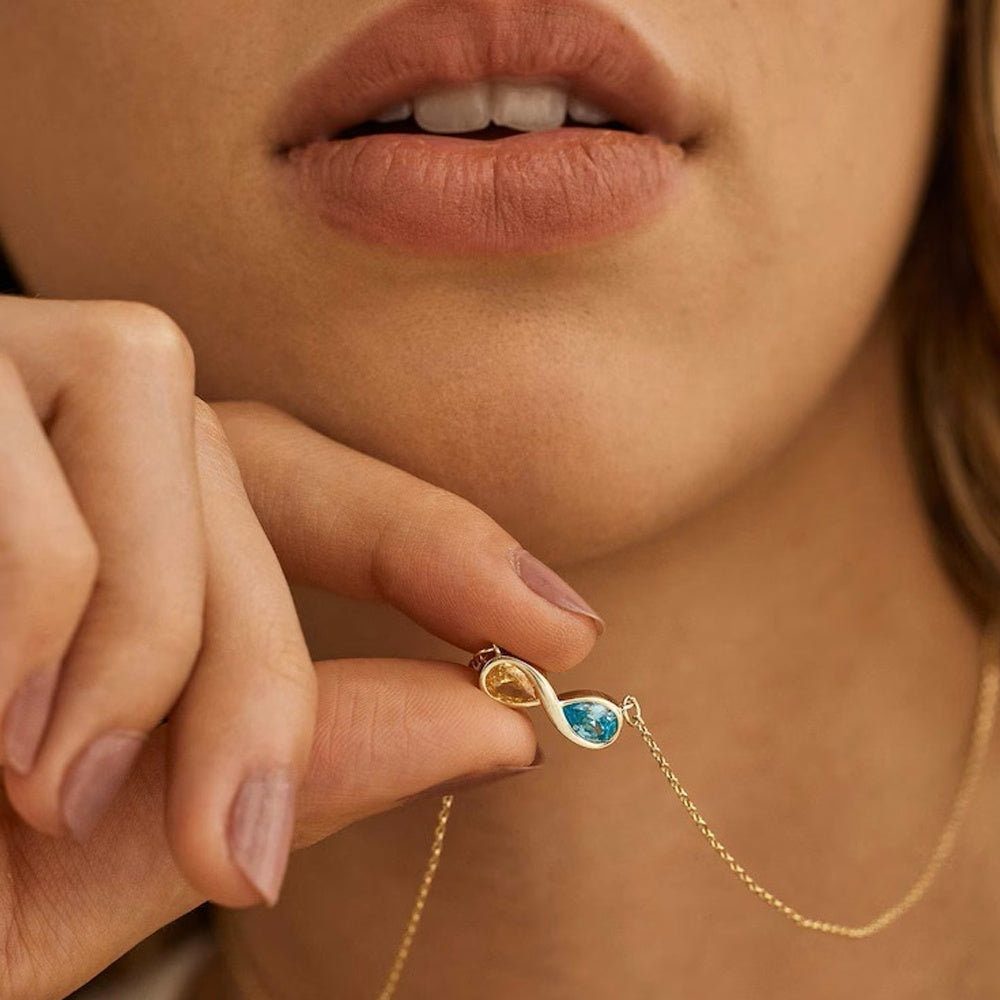 Close-up of a gold infinity birthstone necklace with a blue gemstone held by a hand near a person's face.