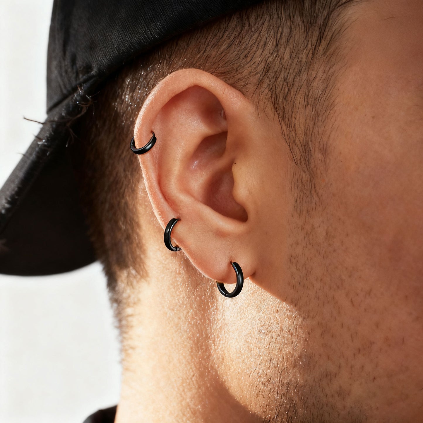 Close-up of a person's ear wearing three black hoop piercings, including a lobe hoop and two cartilage hoop piercings, with a black cap visible at the top.