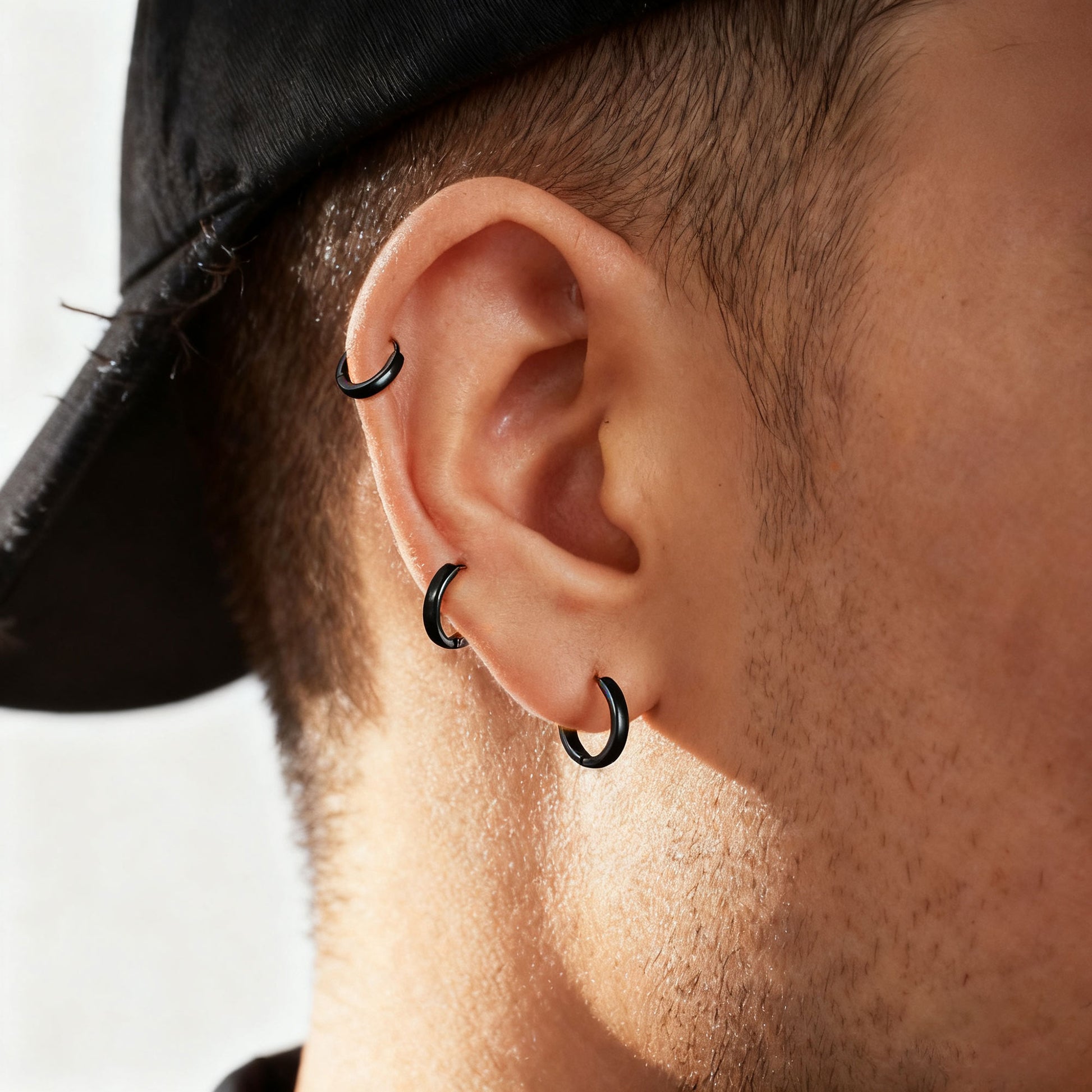 Close-up of a person's ear wearing three black hoop piercings, including a lobe hoop and two cartilage hoop piercings, with a black cap visible at the top.