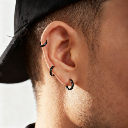 Close-up of a person's ear wearing three black hoop piercings, including a lobe hoop and two cartilage hoop piercings, with a black cap visible at the top.