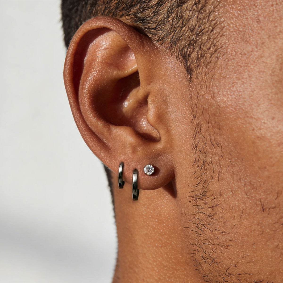 Close-up of a person's ear wearing three earrings: two small huggie earrings and one silver-toned stud with a clear gemstone, against a light neutral background.