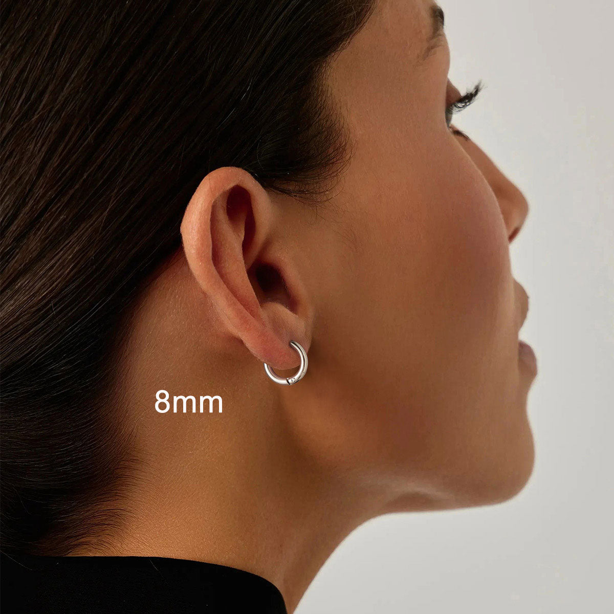 Close-up of an ear wearing a silver huggie earring with '8mm' text on a neutral background