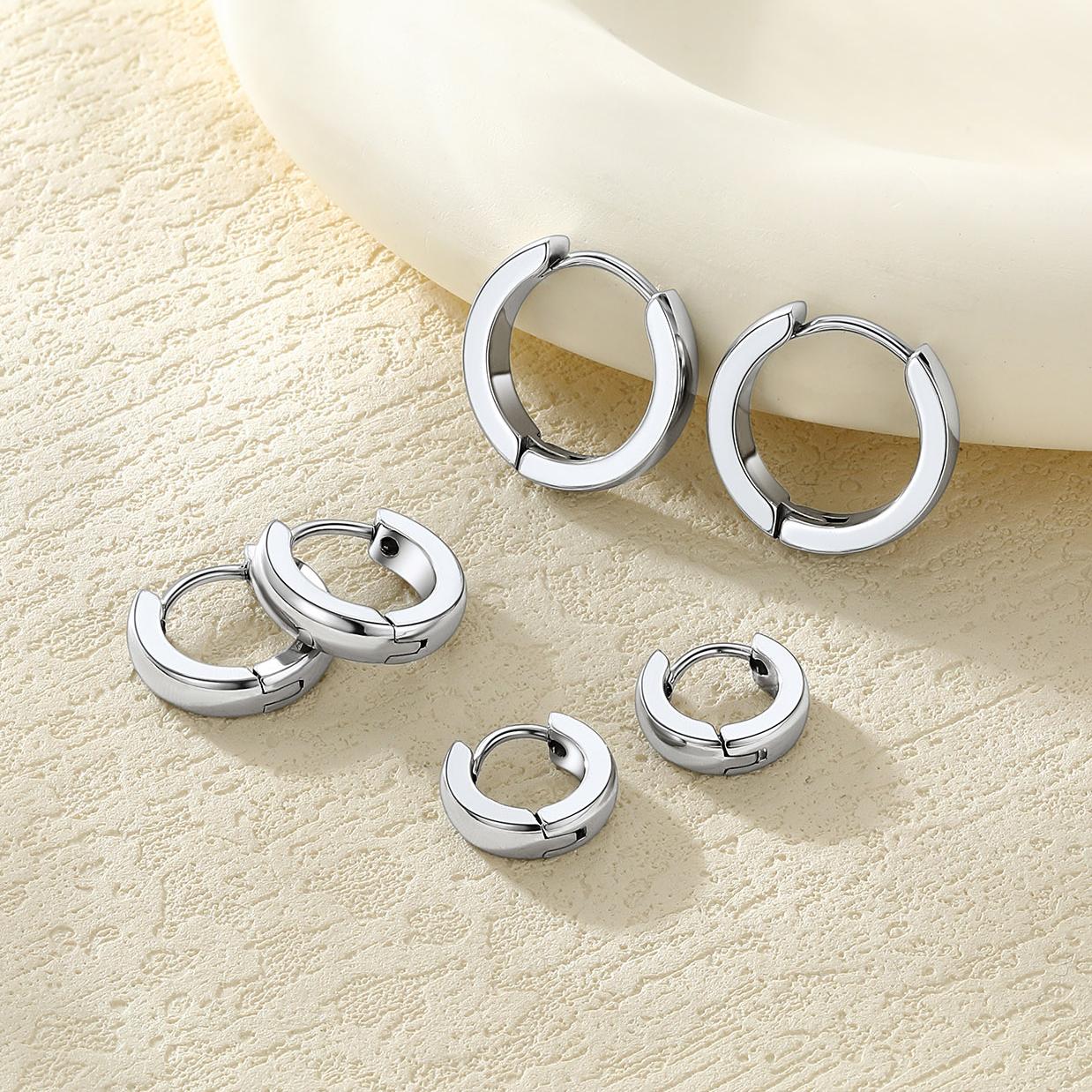 Five pairs of polished silver huggie earrings arranged on a textured beige surface with a white ceramic bowl in the background.