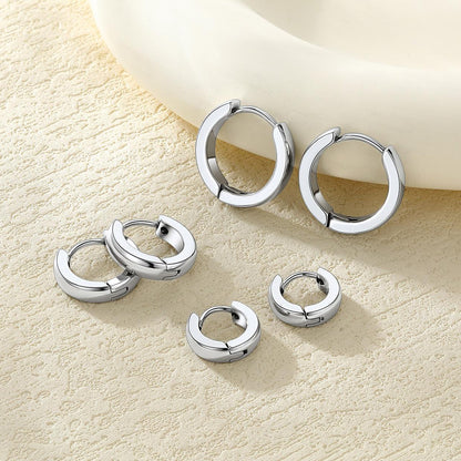 Five pairs of polished silver huggie earrings arranged on a textured beige surface with a white ceramic bowl in the background.