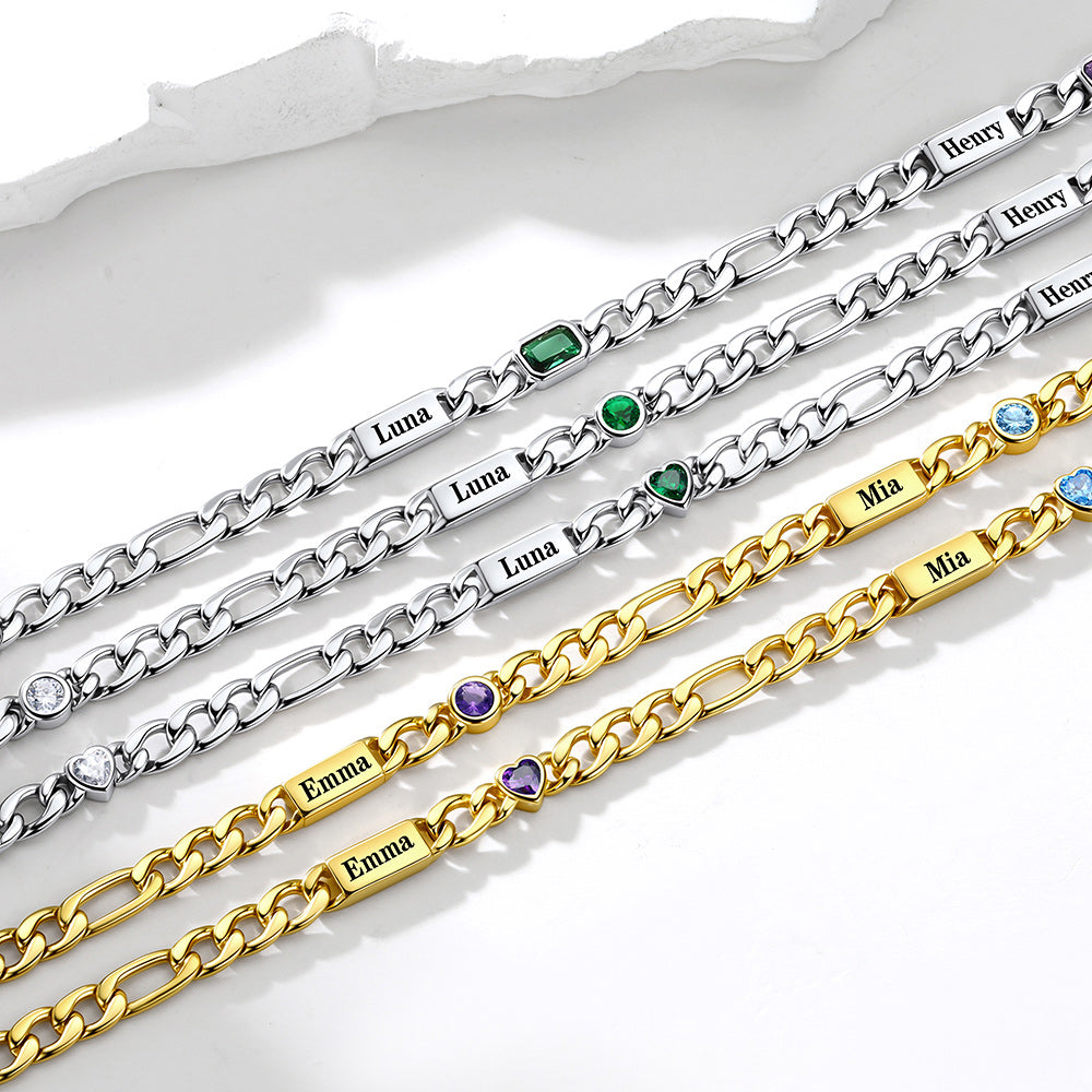 Five personalized name figaro chain necklace in silver and gold tones, each featuring a nameplate engraved with a name (Luna, Henry, Emma, Mia) and small colored gemstone accents, arranged diagonally on a white surface.
