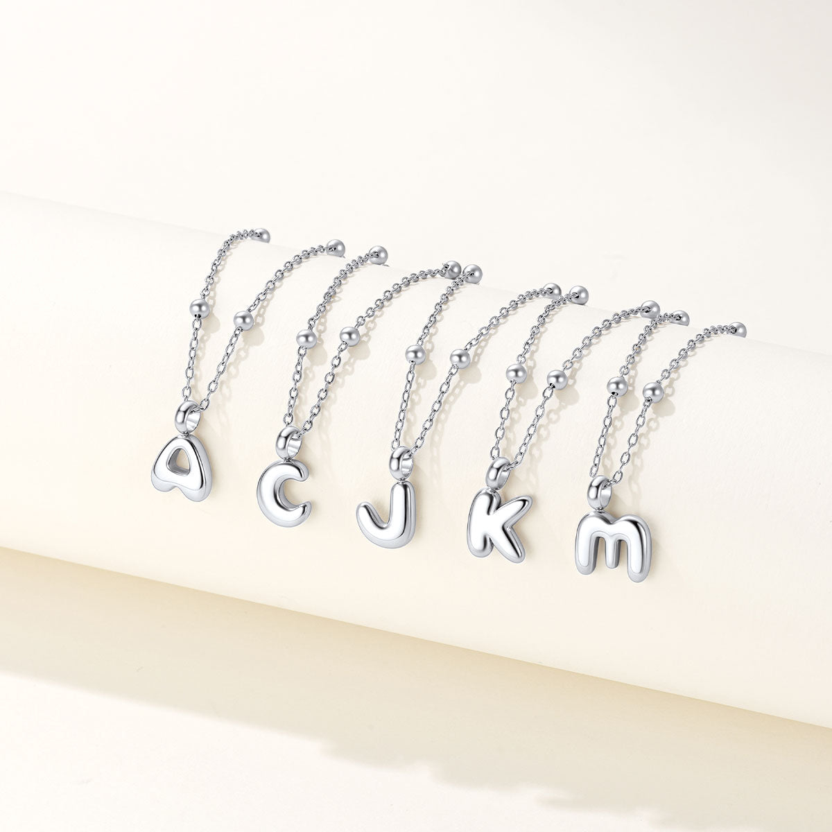 Five silver bubble letter necklaces spelling out 'ACJKM' in lowercase cursive font, arranged in a row on a light cream-colored surface.