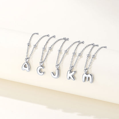 Five silver bubble letter necklaces spelling out 'ACJKM' in lowercase cursive font, arranged in a row on a light cream-colored surface.