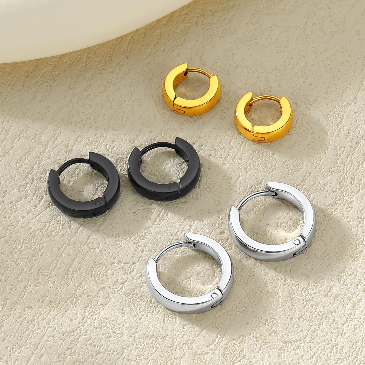 Five small huggie earrings arranged on a textured beige surface, including two gold-tone pairs, two black-tone pairs, and two silver-tone pairs.