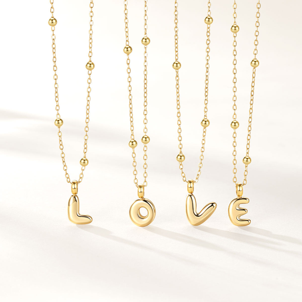 Four gold initial necklaces with bead chains and lowercase letter pendants spelling LOVE, arranged vertically on a clean white background.