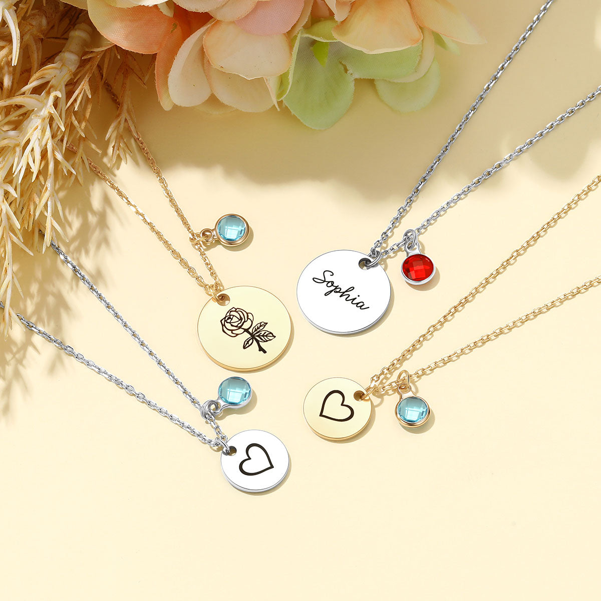 Four personalized round engraved pendant necklaces with chain and small bead charms are arranged on a warm cream background with dried grass and peach flowers. The necklaces include silver and gold chains, silver and gold pendants, and blue and red bead accents.