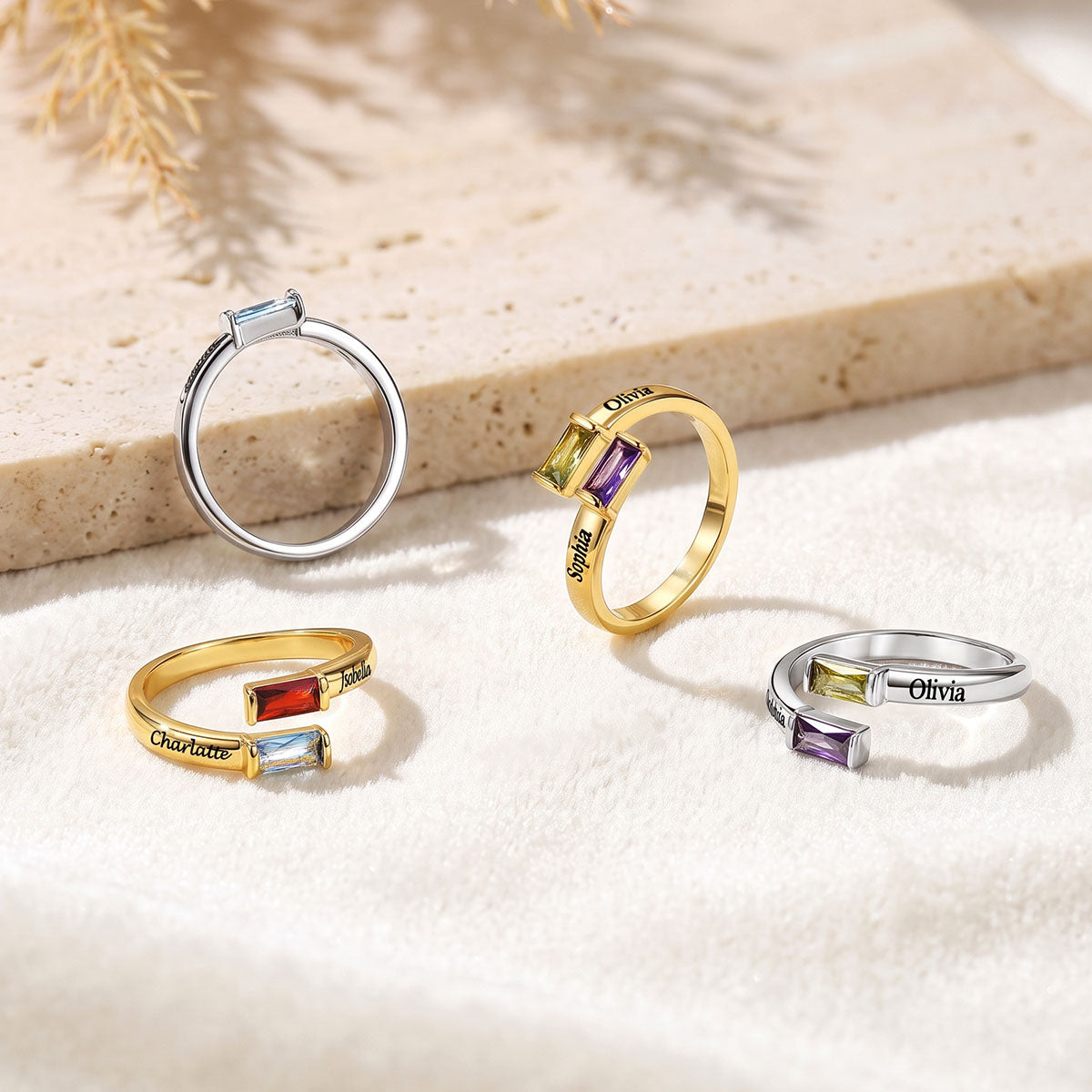 Four personalized silver and gold birthstone rings with rectangular and baguette gemstones, displayed on a textured beige surface with a decorative dried plant in the background.