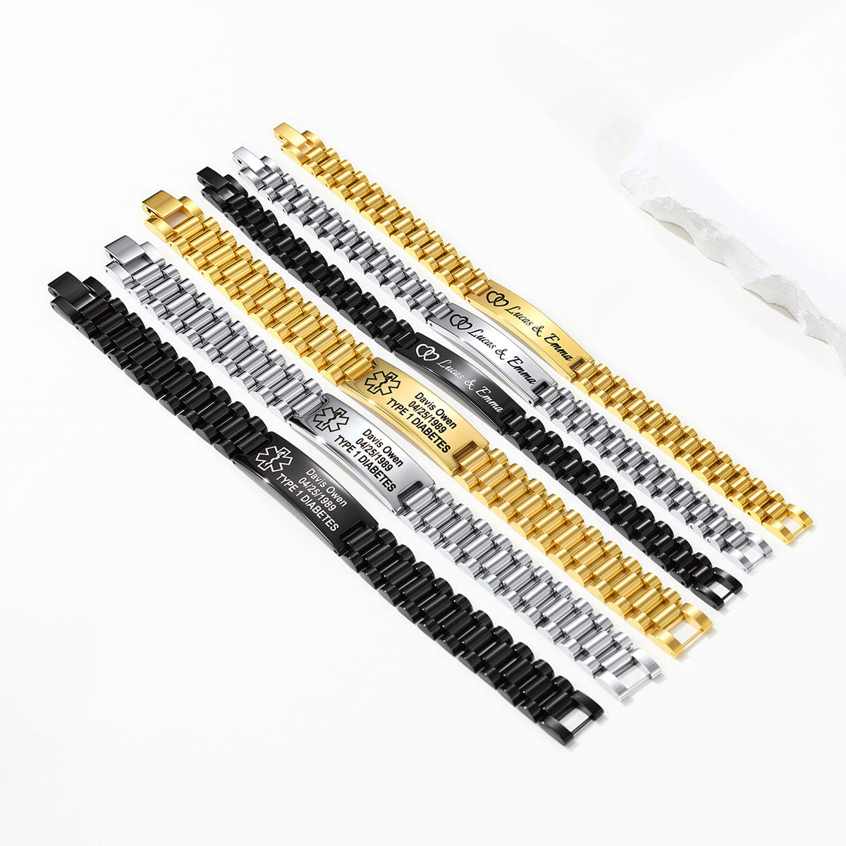 Four personalized stainless steel link Medical Alert bracelets in black, silver, and gold finishes, laid out diagonally on a white background with a small clear plastic bag in the upper right.