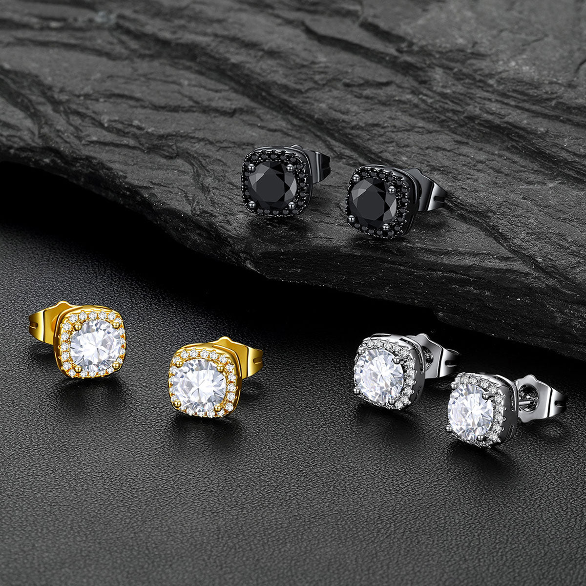 Four square stud earrings displayed on a dark textured surface, including two gold-toned with clear stones, two silver-toned with clear stones, and two black-toned with dark stones. A dark slate slab is placed behind the top pair.