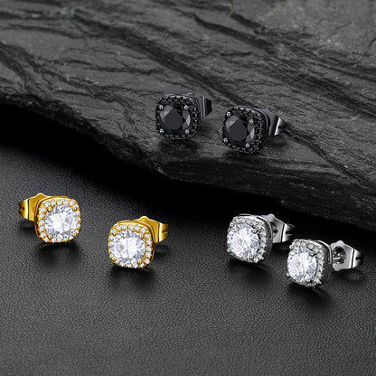 Four square stud earrings displayed on a dark textured surface, including two gold-toned with clear stones, two silver-toned with clear stones, and two black-toned with dark stones. A dark slate slab is placed behind the top pair.