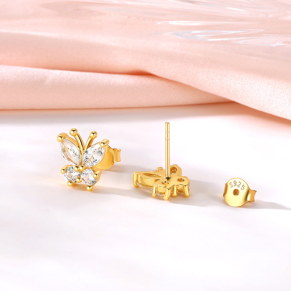 Gold butterfly stud earrings with clear gemstones, shown with one earring facing forward, one showing the back post, and a separate round back clip on a white surface with a soft pink satin background.