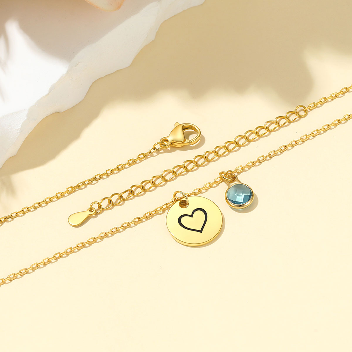 Gold chain anklets with a round gold disc charm necklace featuring an engraved black heart, a small round blue faceted gemstone, and an adjustable lobster clasp, displayed on a warm beige background.