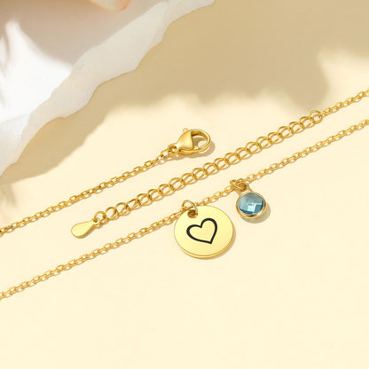 Gold chain anklets with a round gold disc charm necklace featuring an engraved black heart, a small round blue faceted gemstone, and an adjustable lobster clasp, displayed on a warm beige background.