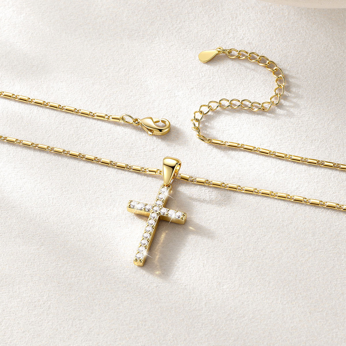 Gold cross necklace with small clear stones, shown on a delicate gold chain with an extender chain on a textured white background.