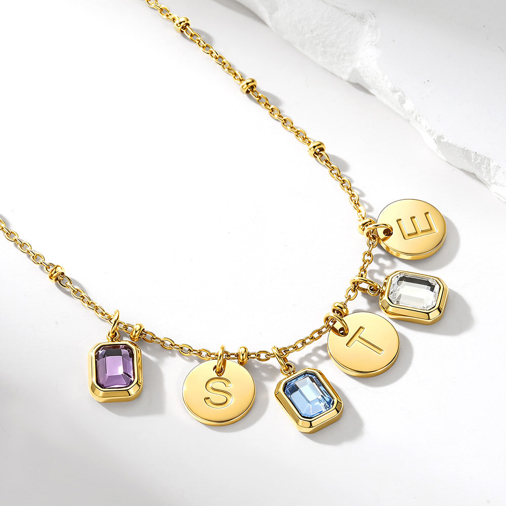 Gold custom birthstone initial necklace with five dangling charms: two rectangular purple and light blue gemstone pendants, two round gold disc pendants engraved with letters, and one clear gemstone pendant, shown on a white background.