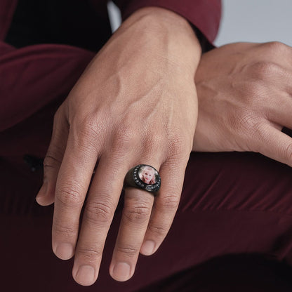 Hand wearing a ring with a small photo on it, against a dark background