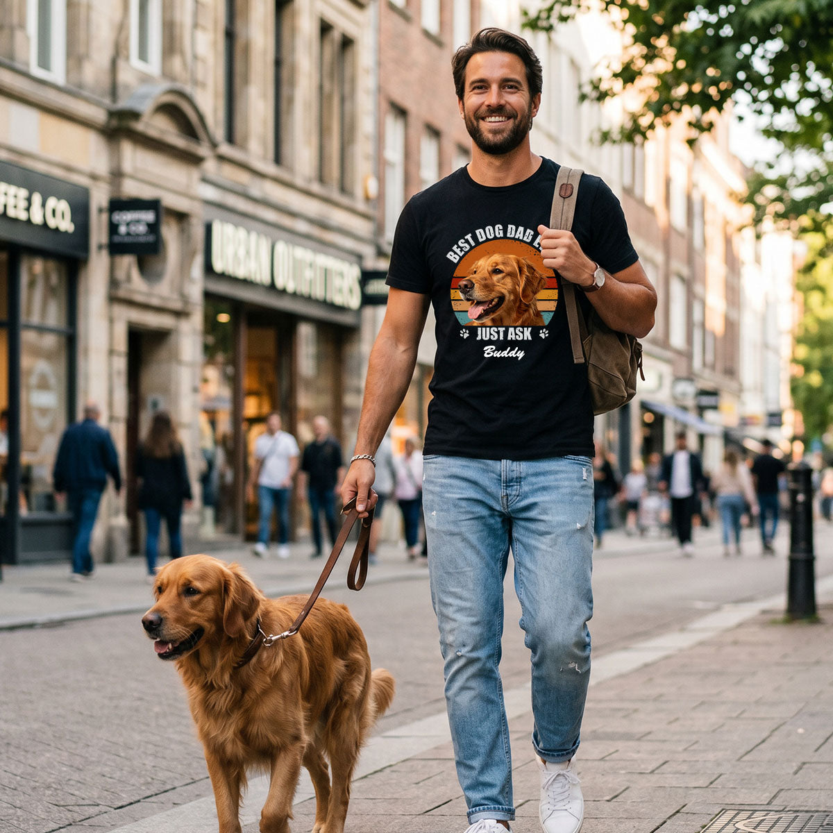 Man walking a dog on a city street wearing a 'Best Dog Dad' t-shirt.