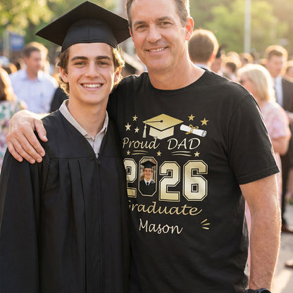 Man wearing a 'Proud Dad of Class of 2026' t-shirt with a photo of a graduate, standing outdoors with people in the background.