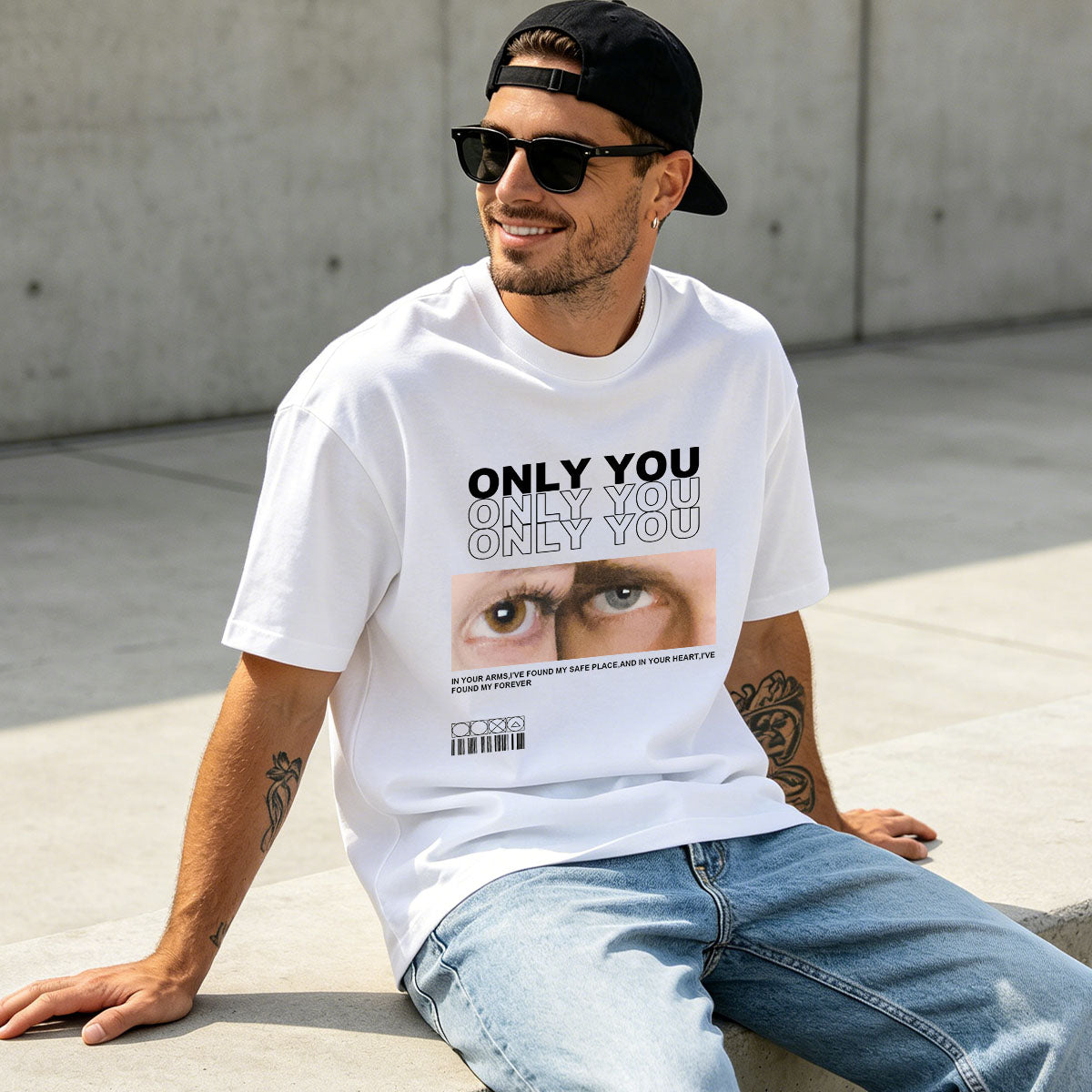 Man wearing a white custom eyes t-shirt with a graphic design and text, sitting on a concrete surface.
