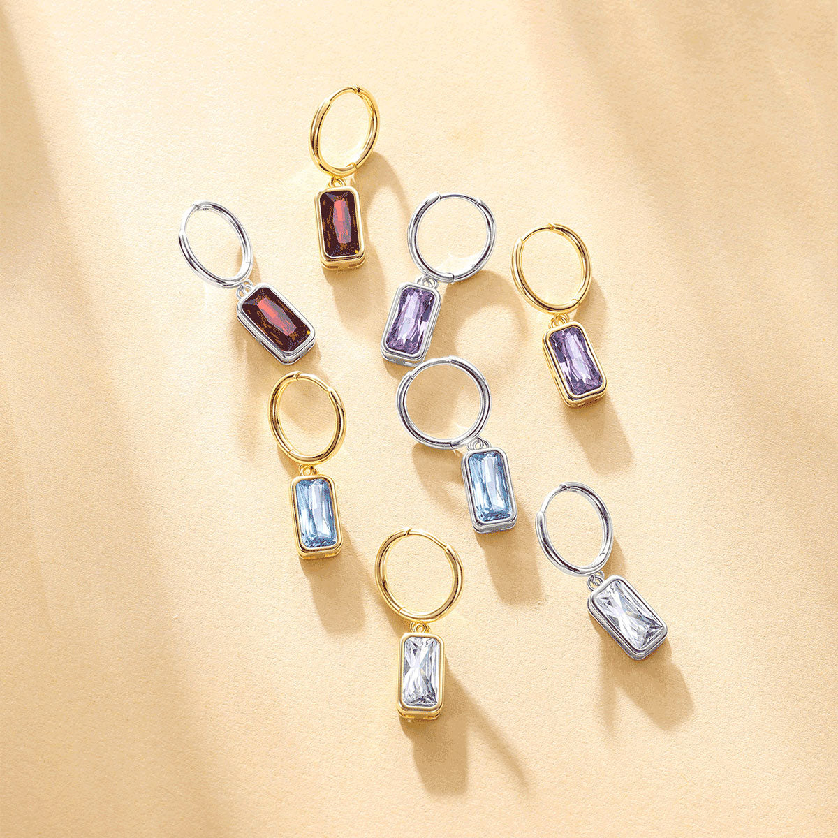 Nine gold and silver hoop earrings with rectangular faceted gemstones in shades of purple, blue, and red, arranged on a warm beige background with soft shadows.