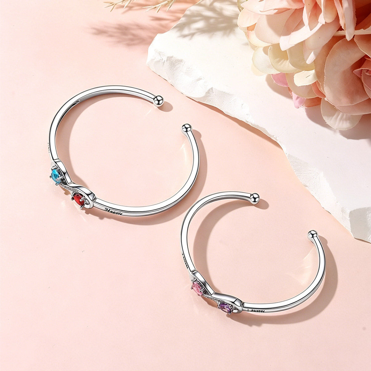 Silver infinity birthstone cuff bracelet with colorful gemstones on a pink background with flowers.