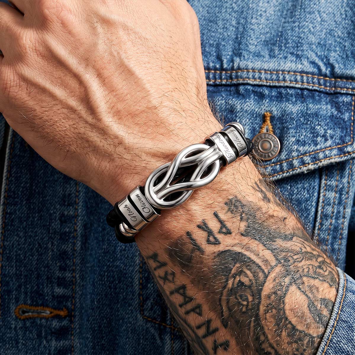 Silver multi name leather bracelet with intricate design worn on a wrist with tattoos, against a denim background.