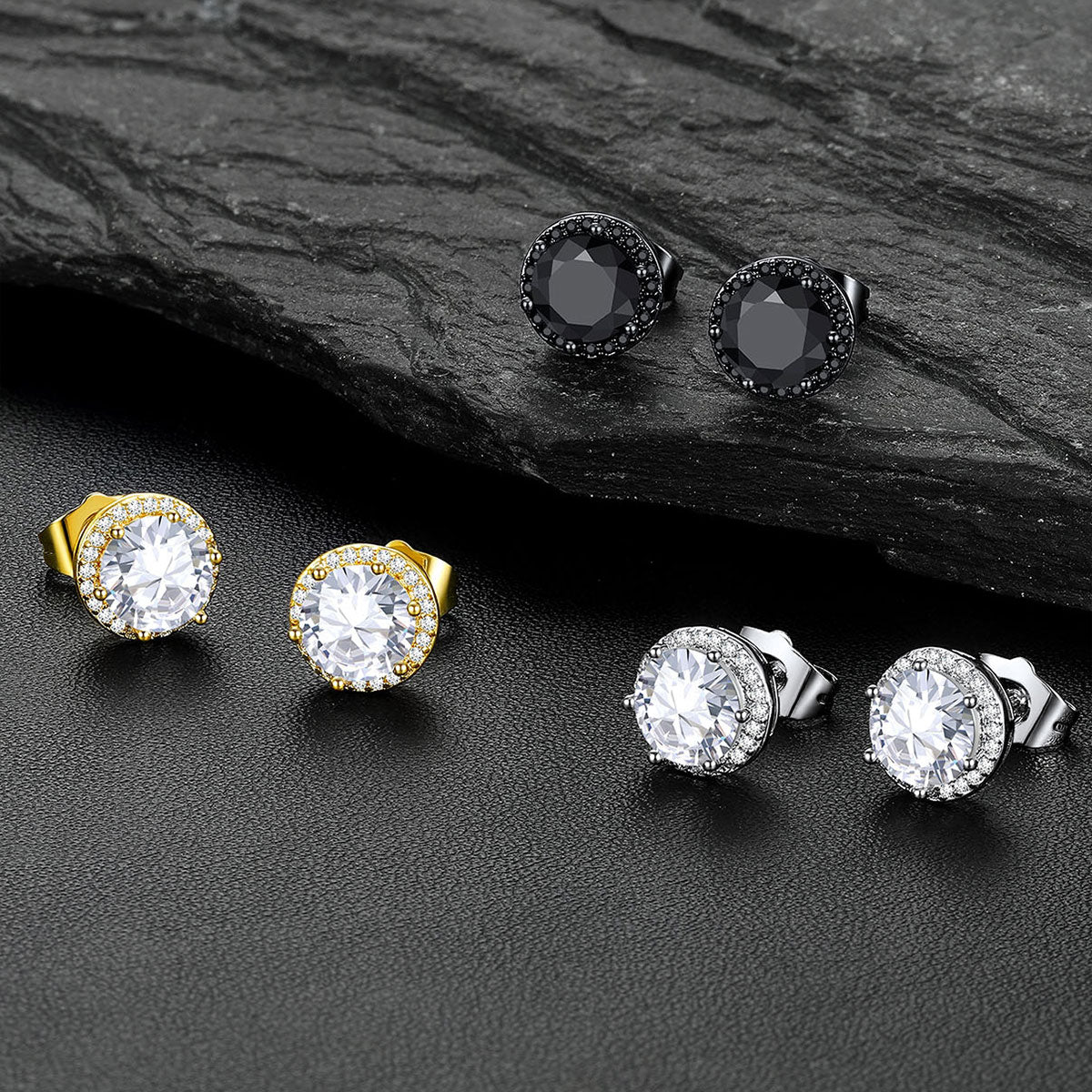 Six pairs of CZ Halo stud earrings arranged on a dark textured surface, including black, yellow gold, silver, and clear stone settings, with a dark slate prop in the background.