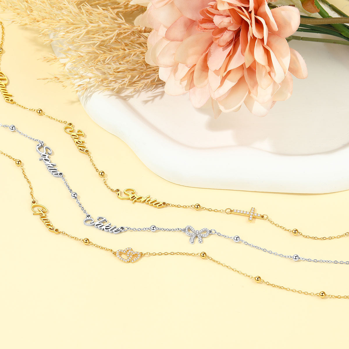 Three delicate nameplate chains in gold, silver, and rose gold tones are arranged on a pale yellow surface with a white dish and a pink flower as styled props.