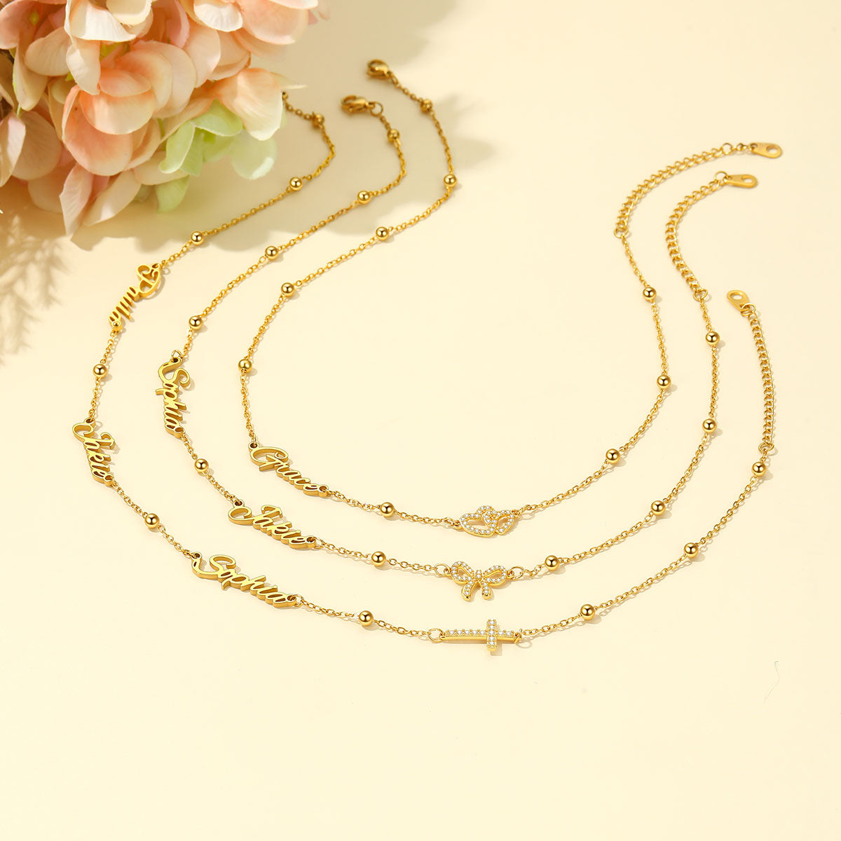 Three gold layered necklaces with script names and a cross pendant, arranged on a light beige background with decorative pink flowers in the top left corner.