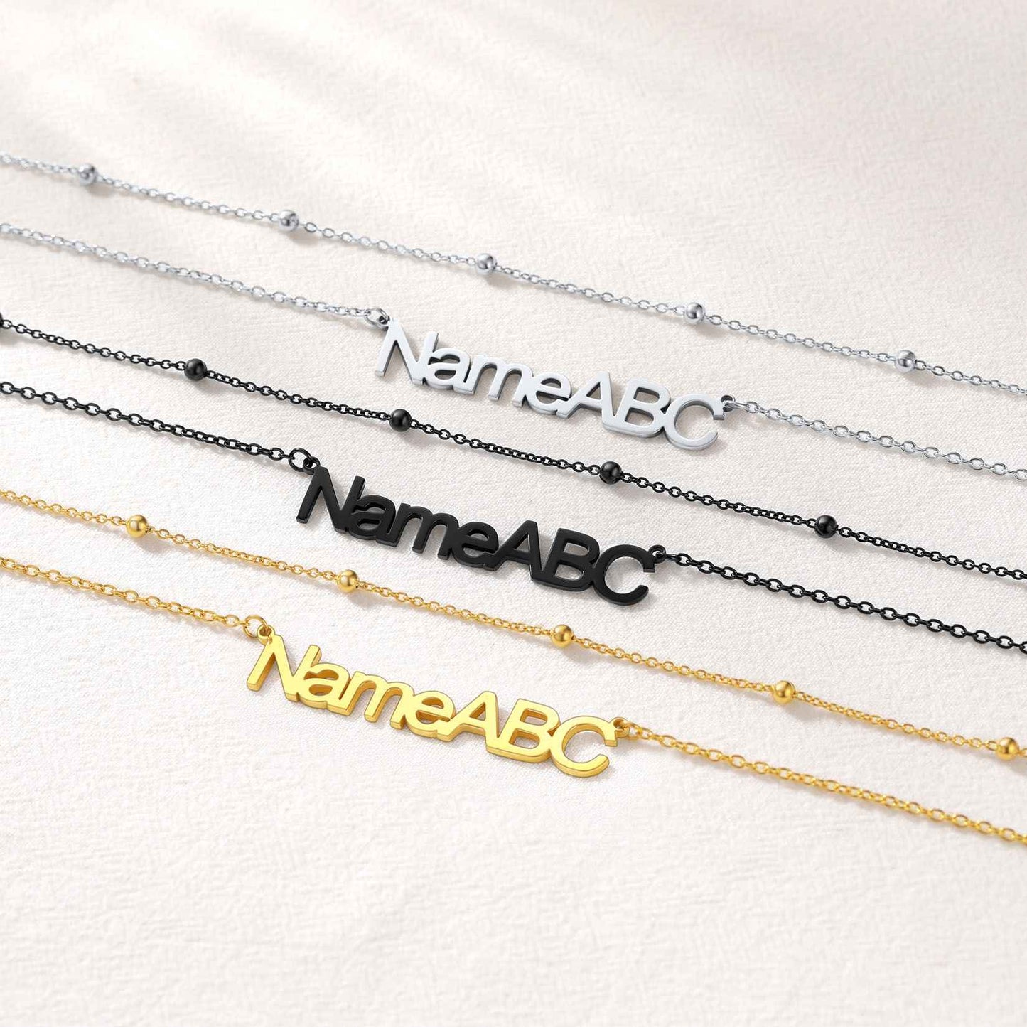Three layered name necklaces with 'NameABC' on them, in silver, black, and gold, on a white background.
