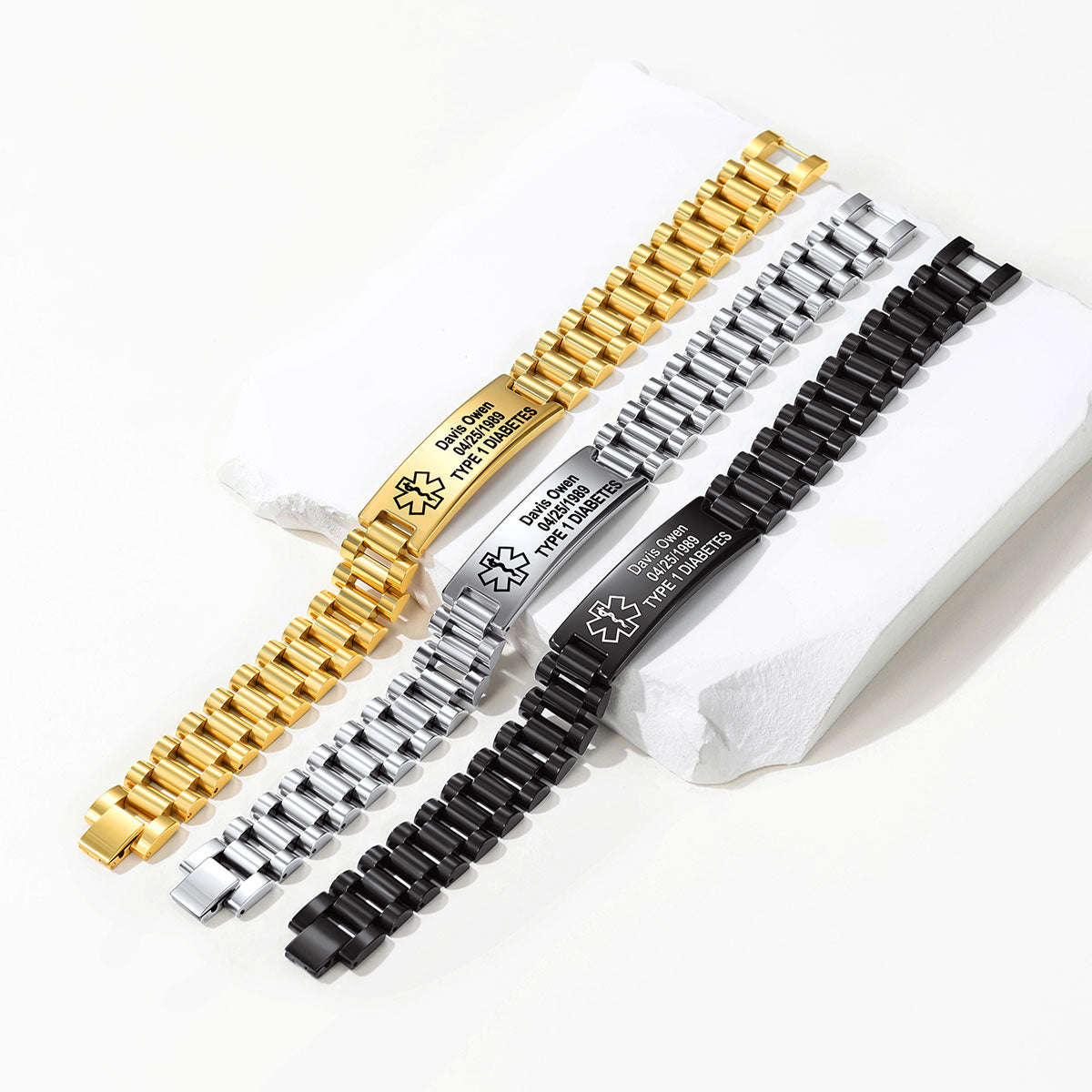 Three metal link ID bracelets in gold, silver, and black finishes, laid diagonally on white display blocks. Each bracelet has a central engraved plate with a monogram and the text 'Distinguished Staff of the.