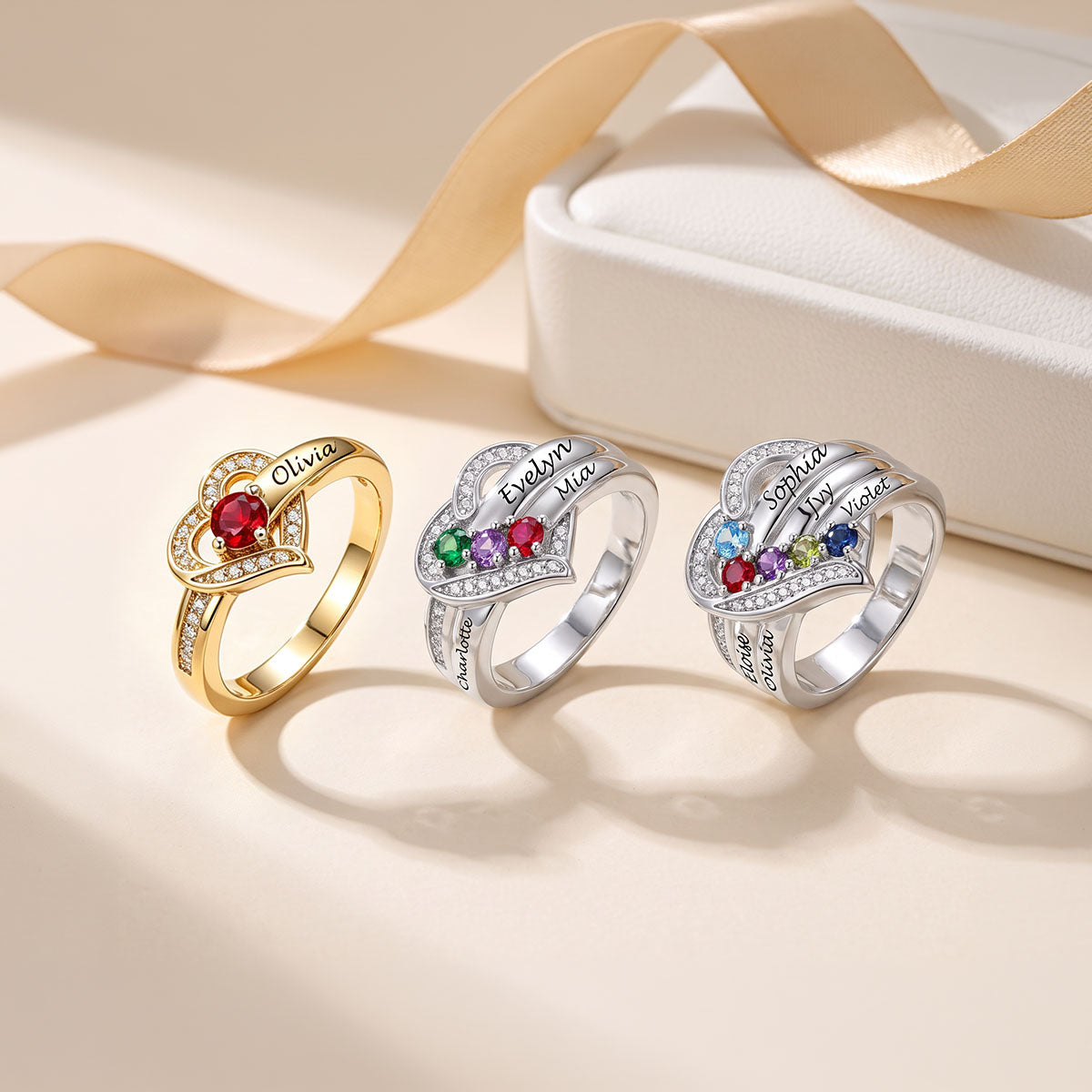 Three personalized heart-shaped rings displayed on a warm beige surface next to a white jewelry box with a gold ribbon. The rings feature custom names engraved on the bands and colorful gemstone accents in red, green, blue, purple, and yellow, with clear pavé stones.