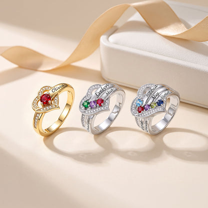 Three personalized heart-shaped rings displayed on a warm beige surface next to a white jewelry box with a gold ribbon. The rings feature custom names engraved on the bands and colorful gemstone accents in red, green, blue, purple, and yellow, with clear pavé stones.