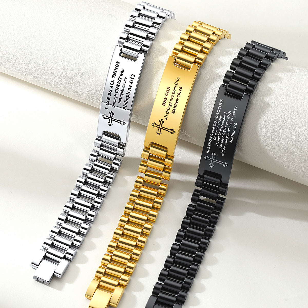 Three personalized stainless steel link bracelets with engraved plates and cross motifs, shown in silver, gold, and black finishes on a white textured background.