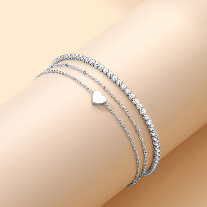 Three silver ankle bracelets set with a heart charm on a beige background