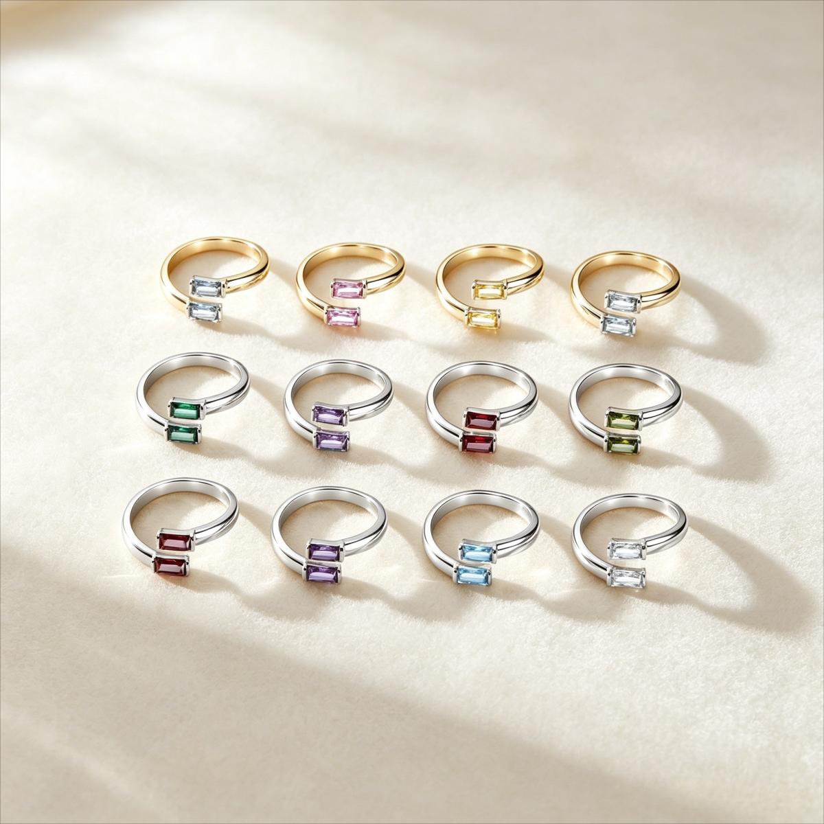 Twelve adjustable split-shank rings arranged in a 3x4 grid on a beige fabric background. The rings feature gold and silver metal bands with small rectangular gemstones in various colors including blue, pink, green, purple, and red.